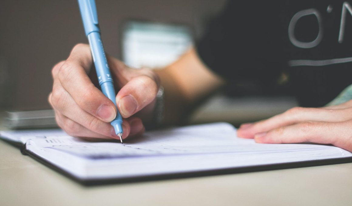 person holding blue ballpoint pen writing in notebook